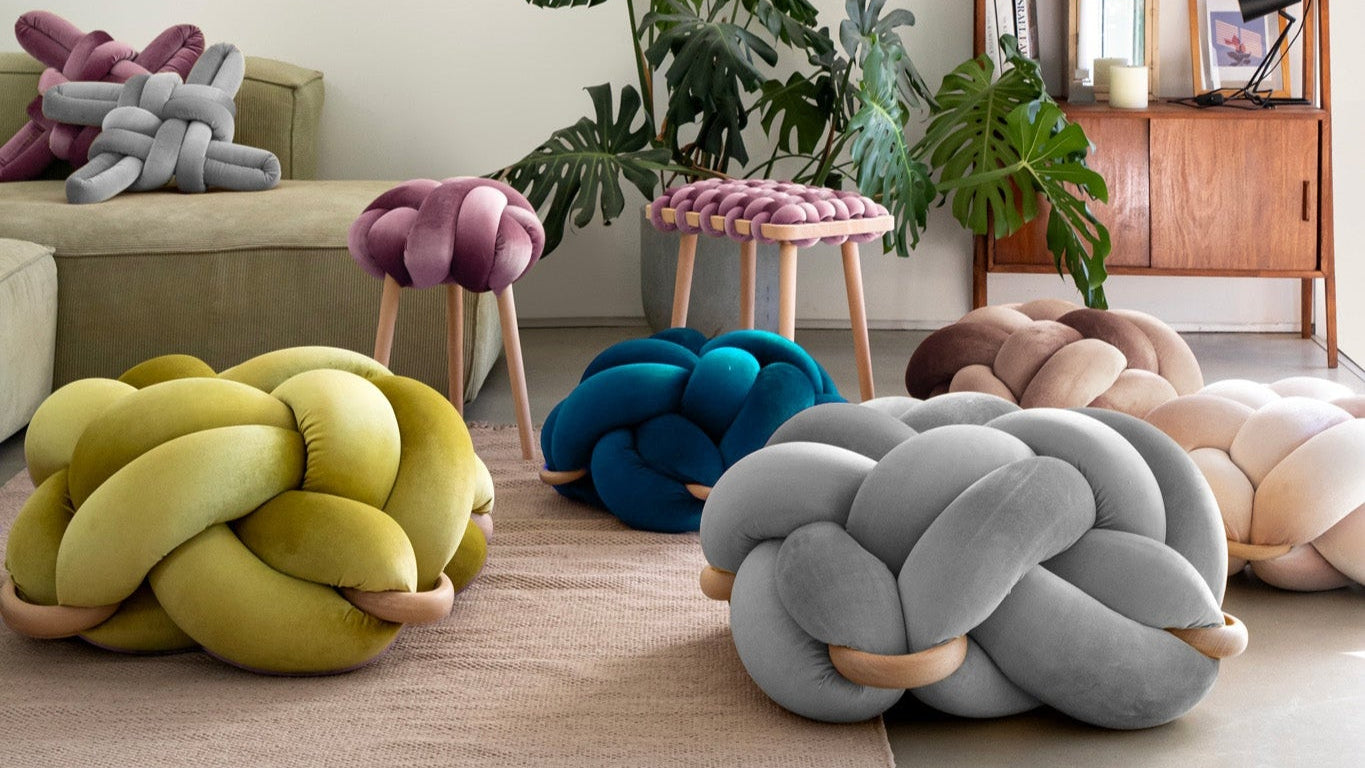 Colorful knot pillows and stools in a living room with plants and a bookshelf.