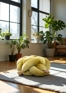 Yellow knotted pillow on a rug in a room with large windows and plants
