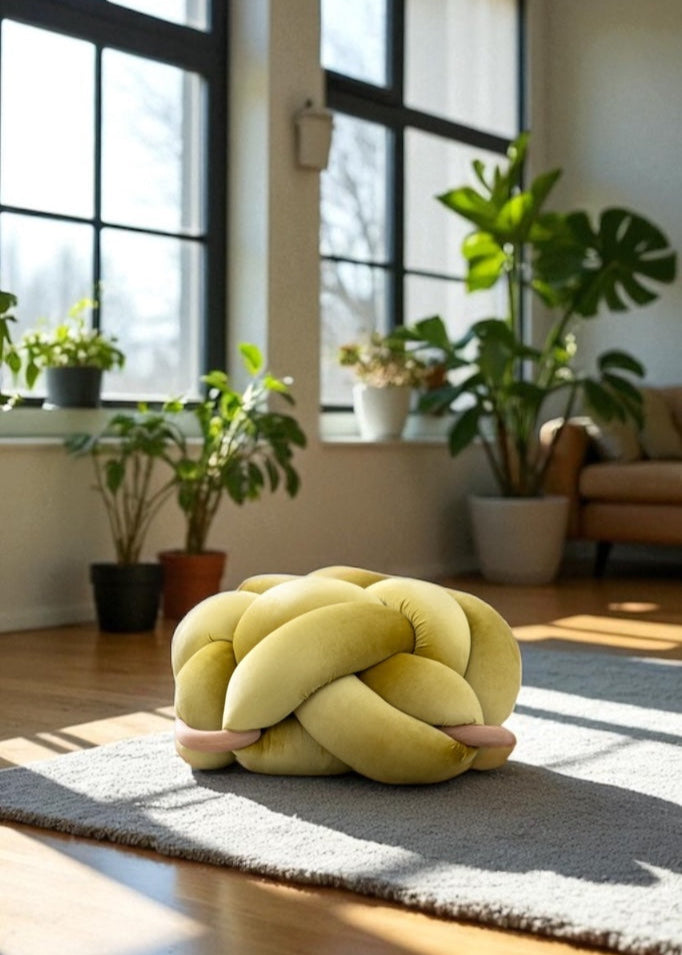 Yellow knotted pillow on a rug in a room with large windows and plants